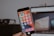 A person is holding a smartphone displaying a screen filled with colorful app icons, mostly in Chinese language. The background is blurred, featuring a computer monitor showing a webpage and a dark object resembling glasses.