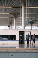 A high-speed train is stationed at a modern railway platform with sleek, structural columns supporting a striped ceiling. Three individuals stand near the train doors, two of them appearing to be engaged in phone calls. The platform has a reflective surface and numbered sections along the edge.
