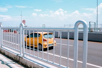 A bright yellow car is driving on a multi-lane road, surrounded by white metal railings. The sky is clear with a few fluffy clouds, and a 'no U-turn' traffic sign is visible in the background.