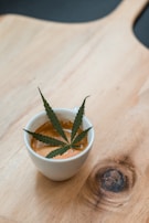 A white cup filled with coffee sits on a wooden surface. A cannabis leaf is placed on top of the coffee as decoration, with a visible knot in the wood next to the cup.