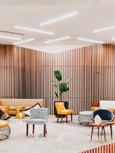 A modern interior space with wooden slat walls and ceiling lighting. The room features a variety of seating options including patterned and solid-colored chairs, a beige couch, and contemporary furniture with geometric designs. A large potted plant adds a touch of greenery to the setting.