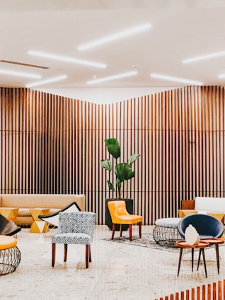 A modern interior space with wooden slat walls and ceiling lighting. The room features a variety of seating options including patterned and solid-colored chairs, a beige couch, and contemporary furniture with geometric designs. A large potted plant adds a touch of greenery to the setting.
