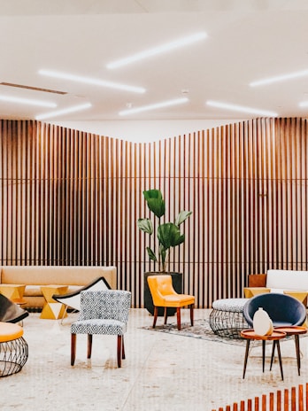 A modern interior space with wooden slat walls and ceiling lighting. The room features a variety of seating options including patterned and solid-colored chairs, a beige couch, and contemporary furniture with geometric designs. A large potted plant adds a touch of greenery to the setting.