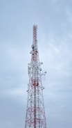 A tall communication tower with a red and white color scheme stands against a cloudy sky. Several antennas and satellite dishes are attached to the structure, indicating its use for broadcasting or telecommunications.