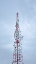 A tall communication tower with a red and white color scheme stands against a cloudy sky. Several antennas and satellite dishes are attached to the structure, indicating its use for broadcasting or telecommunications.