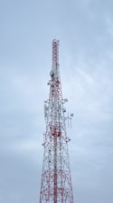 A tall communication tower with a red and white color scheme stands against a cloudy sky. Several antennas and satellite dishes are attached to the structure, indicating its use for broadcasting or telecommunications.
