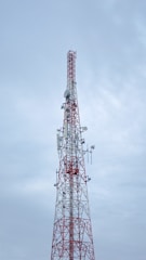A tall communication tower with a red and white color scheme stands against a cloudy sky. Several antennas and satellite dishes are attached to the structure, indicating its use for broadcasting or telecommunications.