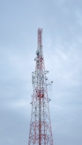 A tall communication tower with a red and white color scheme stands against a cloudy sky. Several antennas and satellite dishes are attached to the structure, indicating its use for broadcasting or telecommunications.