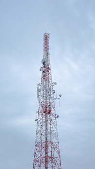 A tall communication tower with a red and white color scheme stands against a cloudy sky. Several antennas and satellite dishes are attached to the structure, indicating its use for broadcasting or telecommunications.