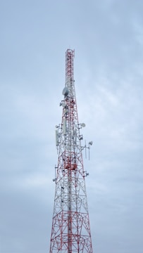 A tall communication tower with a red and white color scheme stands against a cloudy sky. Several antennas and satellite dishes are attached to the structure, indicating its use for broadcasting or telecommunications.