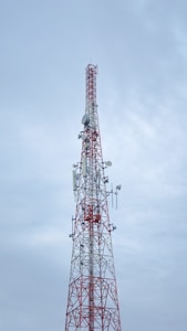 A tall communication tower with a red and white color scheme stands against a cloudy sky. Several antennas and satellite dishes are attached to the structure, indicating its use for broadcasting or telecommunications.