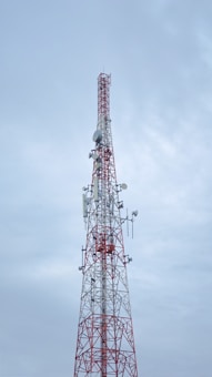 A tall communication tower with a red and white color scheme stands against a cloudy sky. Several antennas and satellite dishes are attached to the structure, indicating its use for broadcasting or telecommunications.