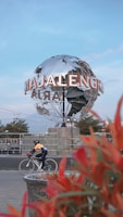 A large metallic globe structure with the word 'Majalengka' prominently displayed stands in an urban setting. A person wearing a helmet and cycling attire rides a road bike in the foreground, and bright red foliage adds a splash of color to the scene. The sky is clear with a light blue hue, suggesting it's a calm day.