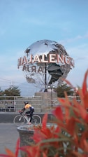A large metallic globe structure with the word 'Majalengka' prominently displayed stands in an urban setting. A person wearing a helmet and cycling attire rides a road bike in the foreground, and bright red foliage adds a splash of color to the scene. The sky is clear with a light blue hue, suggesting it's a calm day.
