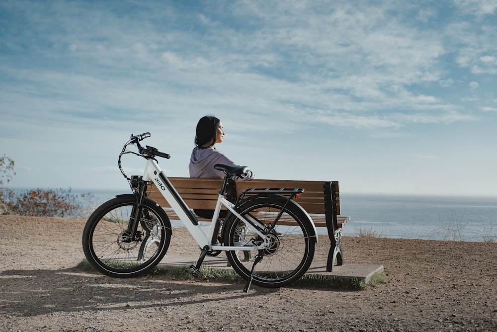 homem na camisa preta sentado no banco de madeira marrom ao lado da bicicleta preta e branca durante o dia