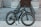 A modern electric bike is positioned on a tiled surface in front of a flight of large, grey stone stairs leading to a building entrance. The bike features thick, durable tires suitable for various terrains and a sleek black frame with an electric motor integrated into the design.