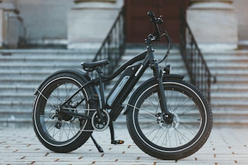 A modern electric bike is positioned on a tiled surface in front of a flight of large, grey stone stairs leading to a building entrance. The bike features thick, durable tires suitable for various terrains and a sleek black frame with an electric motor integrated into the design.