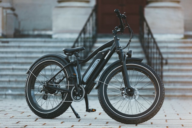 A modern electric bike is positioned on a tiled surface in front of a flight of large, grey stone stairs leading to a building entrance. The bike features thick, durable tires suitable for various terrains and a sleek black frame with an electric motor integrated into the design.