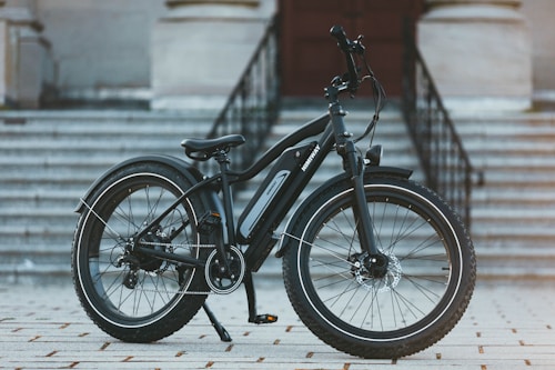 A modern electric bike is positioned on a tiled surface in front of a flight of large, grey stone stairs leading to a building entrance. The bike features thick, durable tires suitable for various terrains and a sleek black frame with an electric motor integrated into the design.