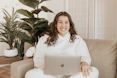A smiling woman sitting at a desk with a laptop, surrounded by plants and cozy decor.
