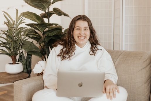 A person is sitting on a beige sofa, smiling and holding a laptop. Several green plants in white pots are placed around, creating a cozy and relaxed ambiance.
