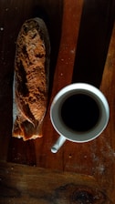 A rustic loaf of bread is placed beside a white cup filled with black coffee on a wooden table, creating a warm and inviting setting.