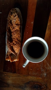 A rustic loaf of bread is placed beside a white cup filled with black coffee on a wooden table, creating a warm and inviting setting.