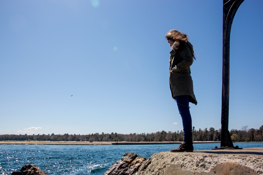 woman in brown jacket standing on rock near body of water during daytime