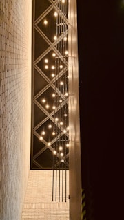 A series of hanging light bulbs are arranged in a vertical pattern against a backdrop of diagonal grid patterns on the ceiling. The walls are made of light-colored bricks, and there is a strong contrast with the darker ceiling.