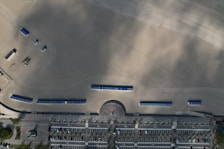 An aerial view of a beach area with rows of blue beach huts aligned neatly on the sand. There's a parking lot in the lower part of the image with marked parking spaces and some parked cars. The sandy area appears to be raked or groomed, showcasing lined patterns. A circular structure or base is present near the top left.