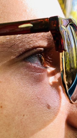 A close-up of a human eye, partially covered by the frame of a pair of sunglasses. The glasses have a thick, dark frame with a slight tortoiseshell pattern. The sunlight casts a sharp shadow on the skin, highlighting the textured surface and individual hairs around the eye.