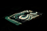 A pattern on a carpet is illuminated by a shaft of light, creating a striking contrast between the lit area and the surrounding darkness. The design features bold, swirling shapes with hues of green, beige, and white.