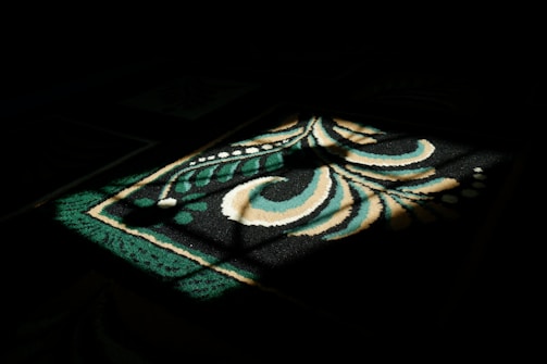 A pattern on a carpet is illuminated by a shaft of light, creating a striking contrast between the lit area and the surrounding darkness. The design features bold, swirling shapes with hues of green, beige, and white.