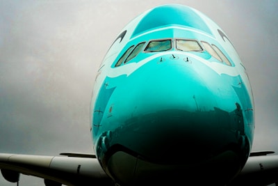 A close-up view of the nose of a turquoise airplane with windows visible at the top. The sky is overcast, creating a dull, gray background that contrasts with the shiny surface of the aircraft.