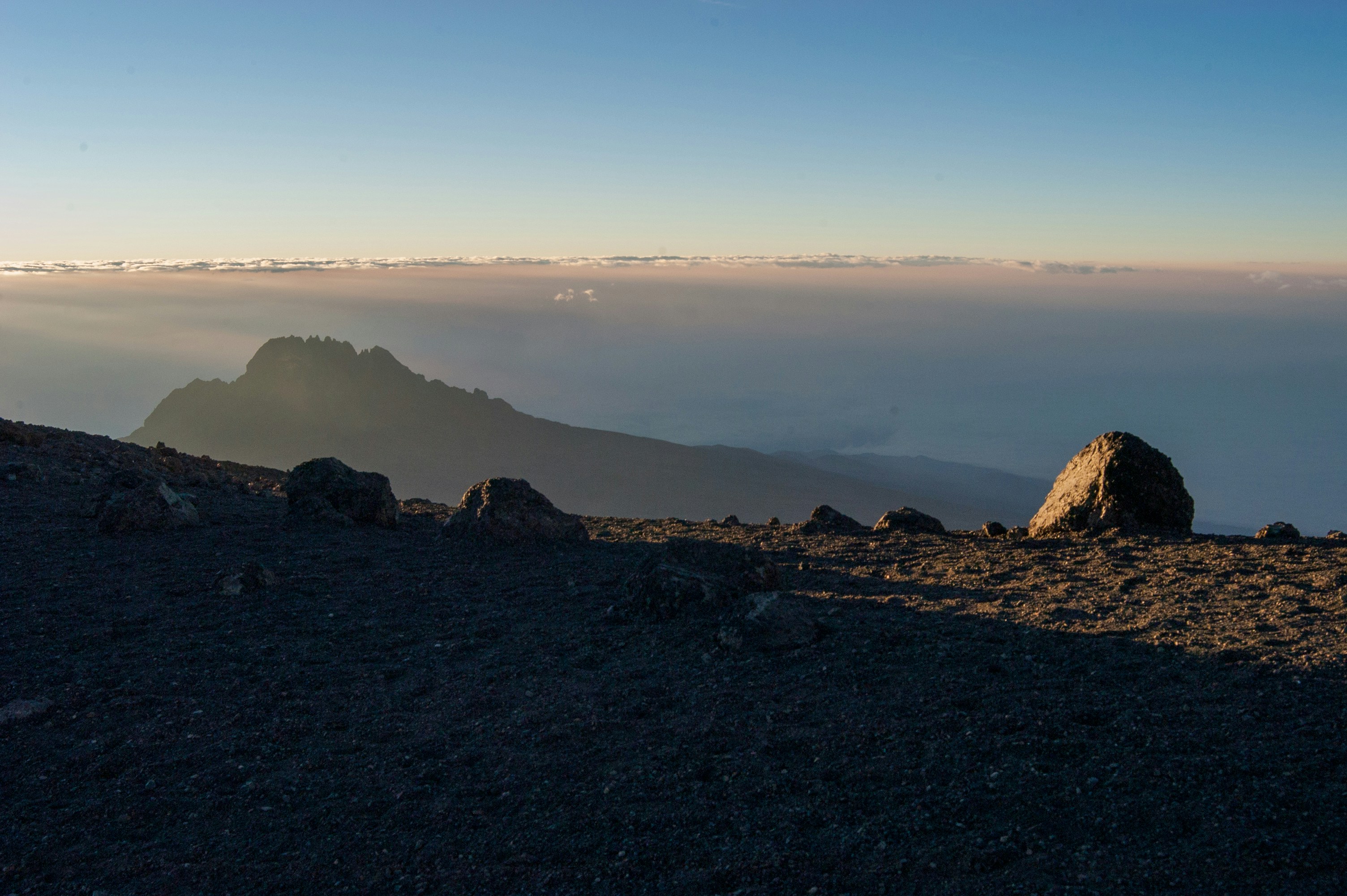 brown rock formation on the sea shore during daytime, The view across the Kilimanjaro Region from the Rongai route. Rocks in the foreground on sand and grit. Mount Mawenzie in view, just. Looks a little like an alien landscape