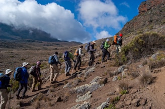 A group of hikers are trekking up a mountain trail surrounded by rocky terrain and patches of vegetation under a partly cloudy sky.