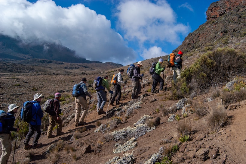 A group of hikers are trekking up a mountain trail surrounded by rocky terrain and patches of vegetation under a partly cloudy sky.