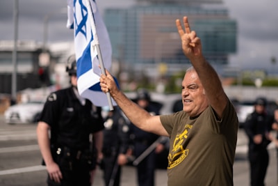 A man in a green shirt raises one arm to make a peace sign while holding the Israeli flag in the other hand. Behind him, several police officers stand on a street. The background includes blurred buildings and vehicles, suggesting a city environment.