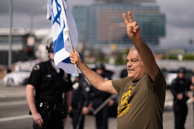 A man in a green shirt raises one arm to make a peace sign while holding the Israeli flag in the other hand. Behind him, several police officers stand on a street. The background includes blurred buildings and vehicles, suggesting a city environment.