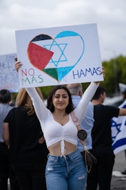 A person holding a protest sign that features a heart design with an Israeli flag motif and the words 'No Más Hamas.' The individual is standing among a group of people, some of whom are carrying additional signs and an Israeli flag.