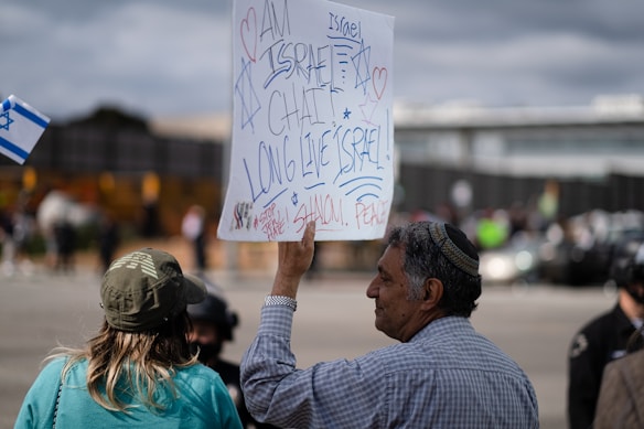 A group of people is standing outdoors holding signs with messages supporting Israel. The signs have handwritten text and symbols such as hearts and the Star of David. The background shows a cloudy sky and a blurred view of other individuals and structures.