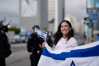 A smiling woman holding an Israeli flag stands outdoors with police officers in the background, in an urban environment with high-rise buildings.