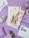 A spiral notebook styled with vintage Parisian fashion-themed prints is placed on a purple surface. It is surrounded by decorative items including gold heart-shaped paper clips, an open pair of scissors, a small bunch of eucalyptus leaves, a white rose, envelopes with vintage stamps, and colored pencils. The background suggests a creative and artistic setting.