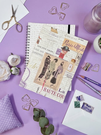 A spiral notebook styled with vintage Parisian fashion-themed prints is placed on a purple surface. It is surrounded by decorative items including gold heart-shaped paper clips, an open pair of scissors, a small bunch of eucalyptus leaves, a white rose, envelopes with vintage stamps, and colored pencils. The background suggests a creative and artistic setting.