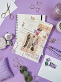 A spiral notebook styled with vintage Parisian fashion-themed prints is placed on a purple surface. It is surrounded by decorative items including gold heart-shaped paper clips, an open pair of scissors, a small bunch of eucalyptus leaves, a white rose, envelopes with vintage stamps, and colored pencils. The background suggests a creative and artistic setting.