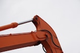 A close-up view of an orange excavator arm with visible hydraulic components against a cloudy sky.