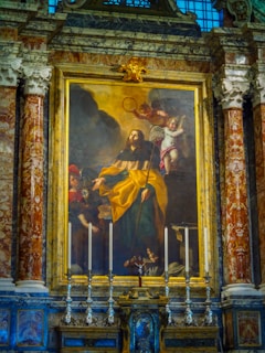 An elaborately decorated altar featuring a large religious painting. The central figure in the artwork is a man wearing a long robe, appearing to hold a staff. A cherub is depicted to the right of this figure, holding a crown or halo. The scene is surrounded by intricate carvings, marble pillars, and several ornate candlesticks arranged symmetrically. The setting suggests a historical or religious significance.