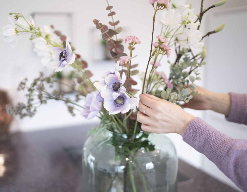 person holding white and purple flowers in clear glass vase