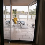 A covered outdoor patio with a view of the neighborhood. There are two black metal chairs with cushions, one with a bright yellow fluffy cushion. Two stools or tables are positioned in front of the chairs, each covered with blue patterned throws. A potted plant is placed on the tiled floor, under a wooden ceiling. Glass railings enclose the area, and trees are visible beyond the patio, under an overcast sky.