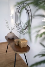 A narrow wooden console table stands against a white wall, adorned with a round decorative wall mirror with a wireframe design. On the table, there is a simple, elegant vase with thin branches and small leaves, alongside two woven baskets. The room has a minimalist, modern aesthetic with a touch of natural elements.